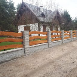 Serwisbruk.pl - Nowoczesne ogrodzenie z drewnianymi przęsłami o falistym kształcie i betonowymi słupkami, na tle domu z ciemnym dachem i zieleni. Widok z zewnątrz.