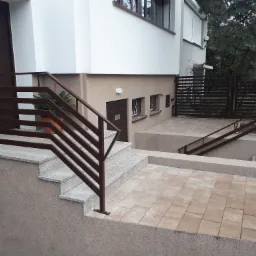 Zewnętrzne schody z szarego granitu prowadzące do budynku, zabezpieczone brązową, poziomą balustradą ze stali nierdzewnej, z widoczną kostką brukową na podwórku i brązową bramą w tle.