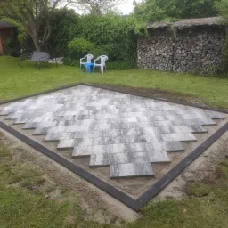 Fragment placu z szarych płyt ułożonych w romboidalny wzór, obramowany czarną kostką, w tle trawnik, drzewa i drewutnia.