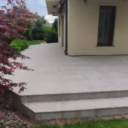 Nowoczesny taras wyłożony jasnymi, kwadratowymi płytami, z dwoma stopniami wejściowymi, obok domu z beżową elewacją i ozdobnym klonem palmowym po lewej stronie.