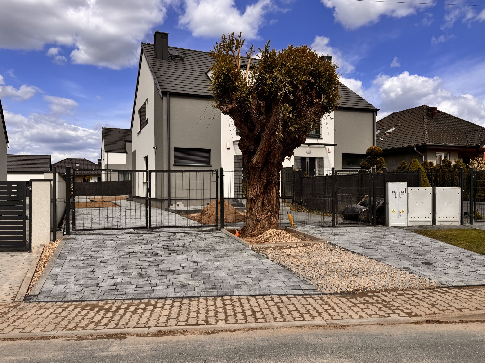 Nowoczesny dom z szarą elewacją i brukowanym podjazdem z metalową bramą. Centralnie umieszczone drzewo dodaje naturalnego charakteru.