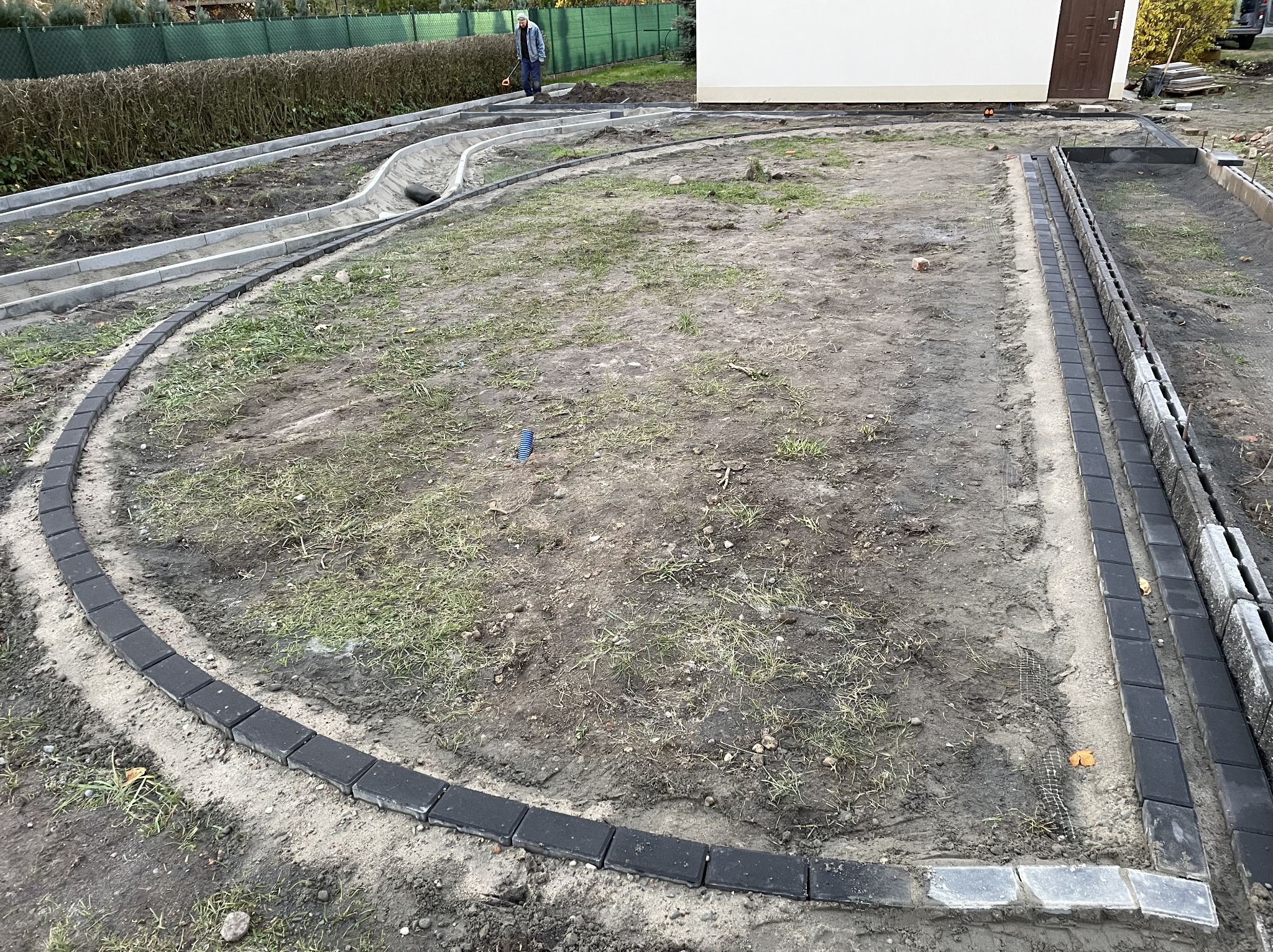 Etap prac: obrzeża z ciemnej kostki ułożone wzdłuż ścieżki i rabaty, przygotowanie terenu pod dalsze prace brukarskie. Widoczny pracownik w tle.