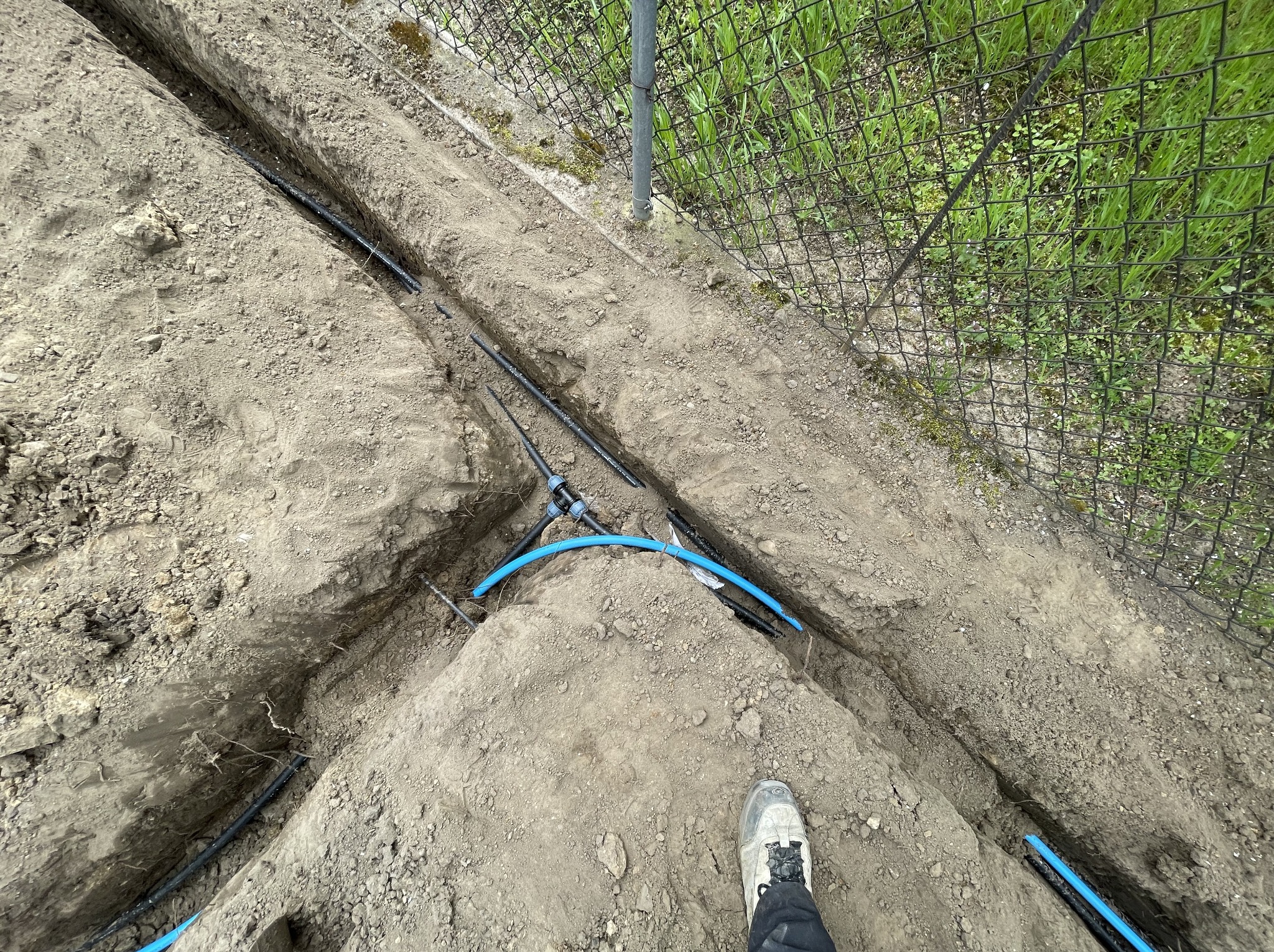 Wykop z ułożonymi rurami nawadniającymi w ziemi, widoczne złącza i fragment buta w prawym dolnym rogu, siatka ogrodzeniowa w tle.