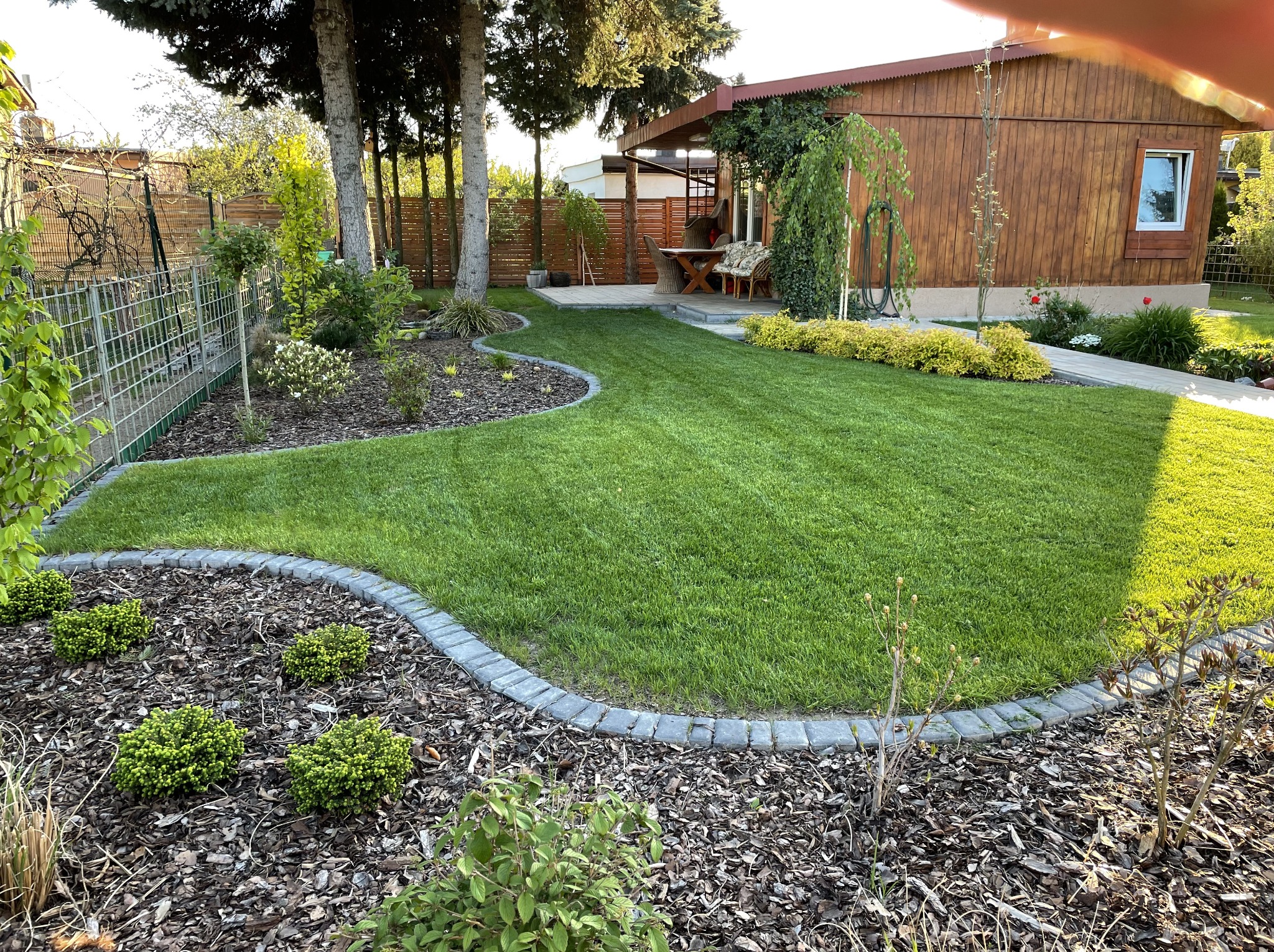 Zadbany ogród z równo przystrzyżonym trawnikiem, obrzeżami z kostki i rabatami z korą. W tle drewniany domek z tarasem i ogrodzenie z desek.