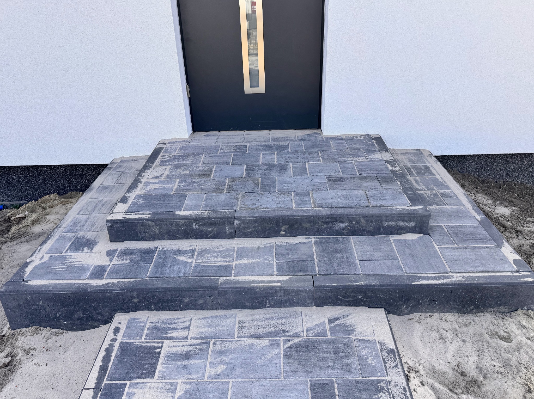 Schody wejściowe z szarej kostki brukowej prowadzące do ciemnych drzwi wejściowych w białym budynku. Widoczne ślady piasku na kostce.