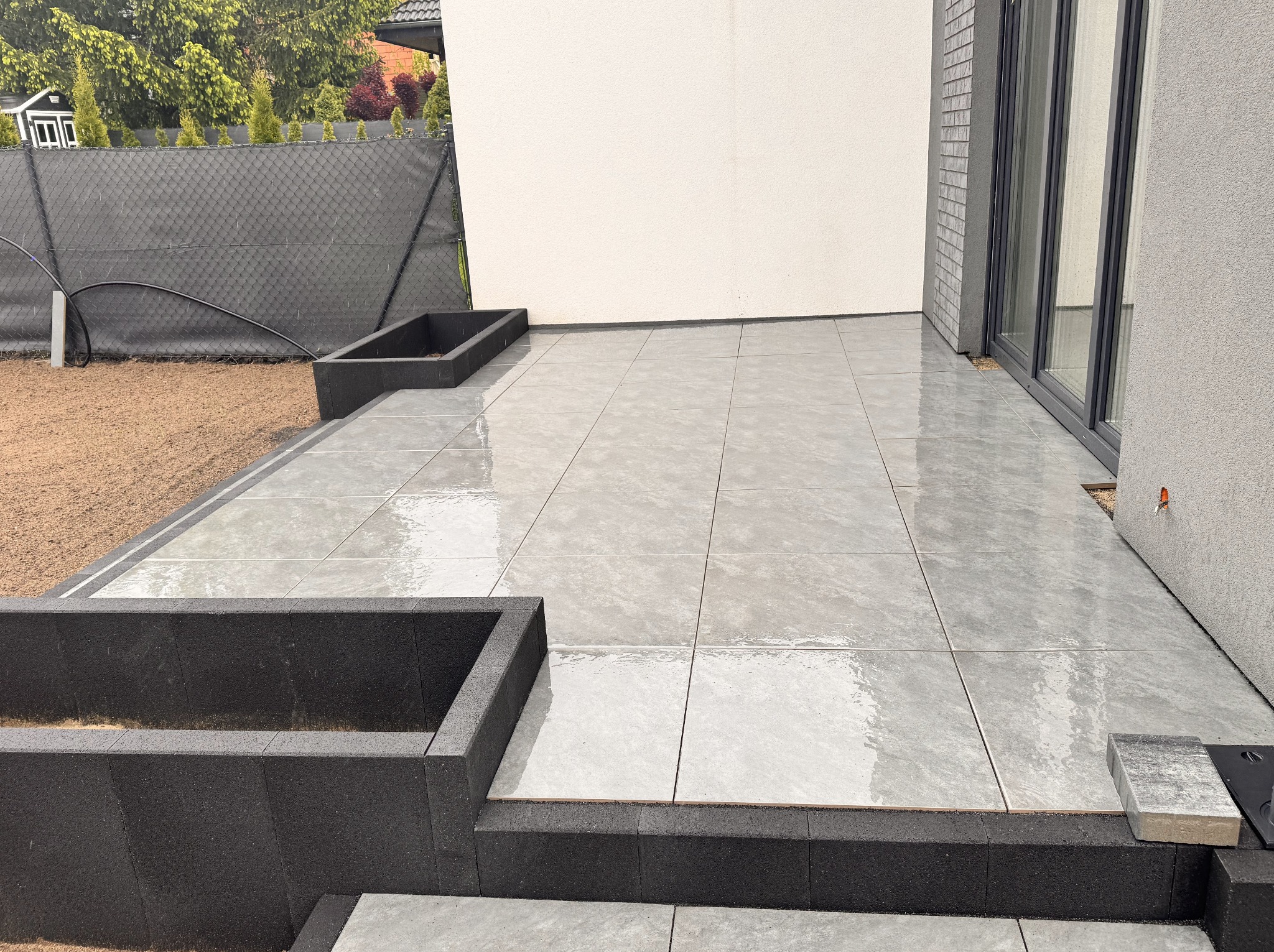 Nowoczesny taras z szarych, kwadratowych płyt, obrzeża z czarnych elementów. Widok na ścianę budynku i fragment ogrodu z czarnym ogrodzeniem.