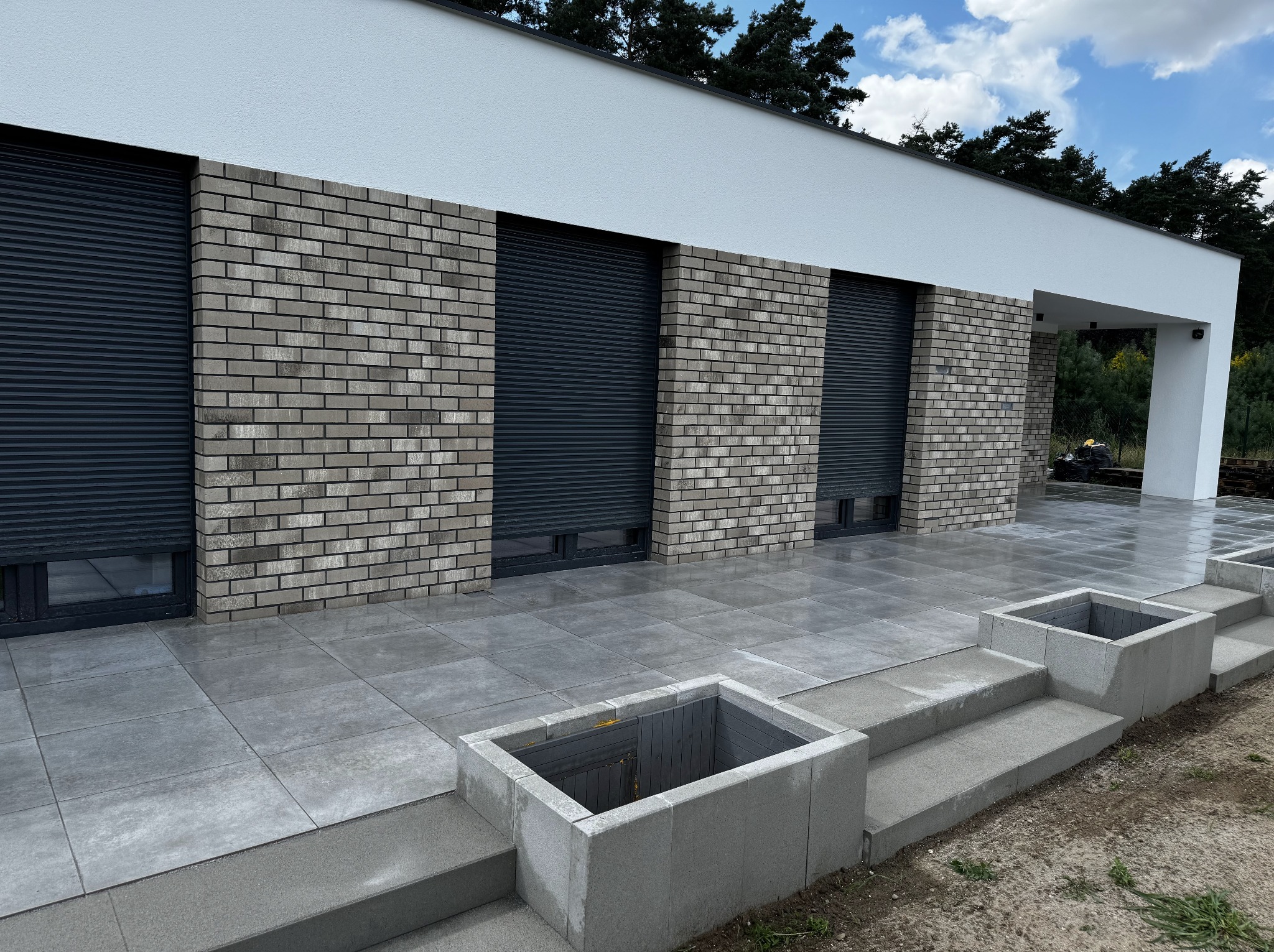 Nowoczesny taras z szarych płyt, zintegrowany ze schodami i donicami z betonu architektonicznego. Elewacja z cegły klinkierowej i rolety zewnętrzne w kolorze antracytowym.