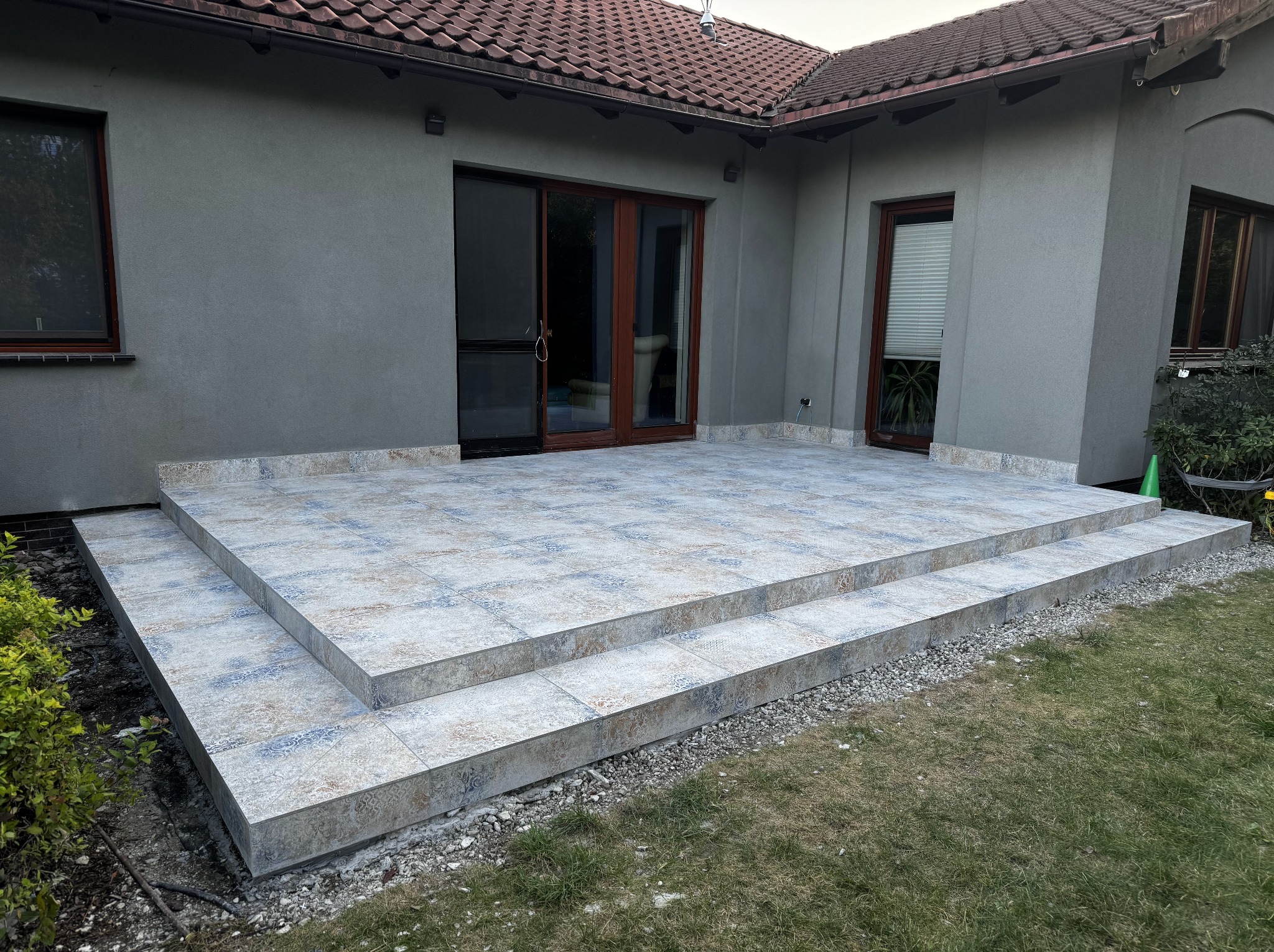 Wykończony taras z płytek ceramicznych z dwoma stopniami, przylegający do szarego budynku z brązowymi oknami i drzwiami. Widok na trawnik i fragment ogrodu.