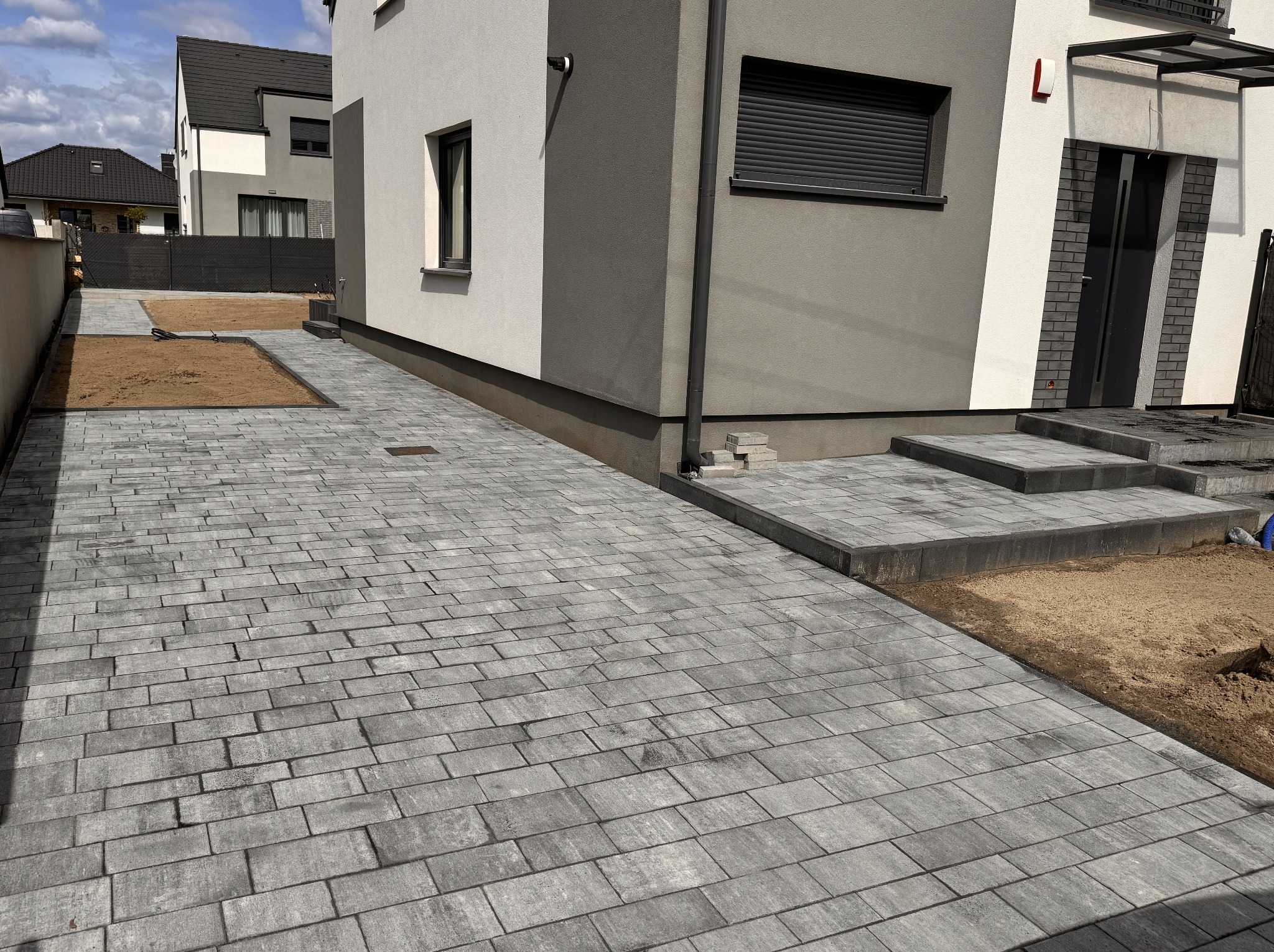 Ułożona szara kostka brukowa prowadząca do nowoczesnego domu z szarymi elementami. Widoczny fragment ogrodu z przygotowanym podłożem pod trawnik.