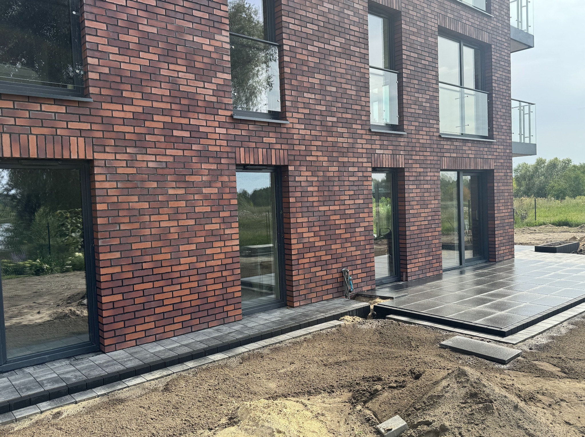 Nowoczesny taras z ciemnej kostki brukowej przy budynku z czerwonej cegły. Widoczne fragmenty ogrodu i balustrady balkonowe. Częściowo ułożona kostka.