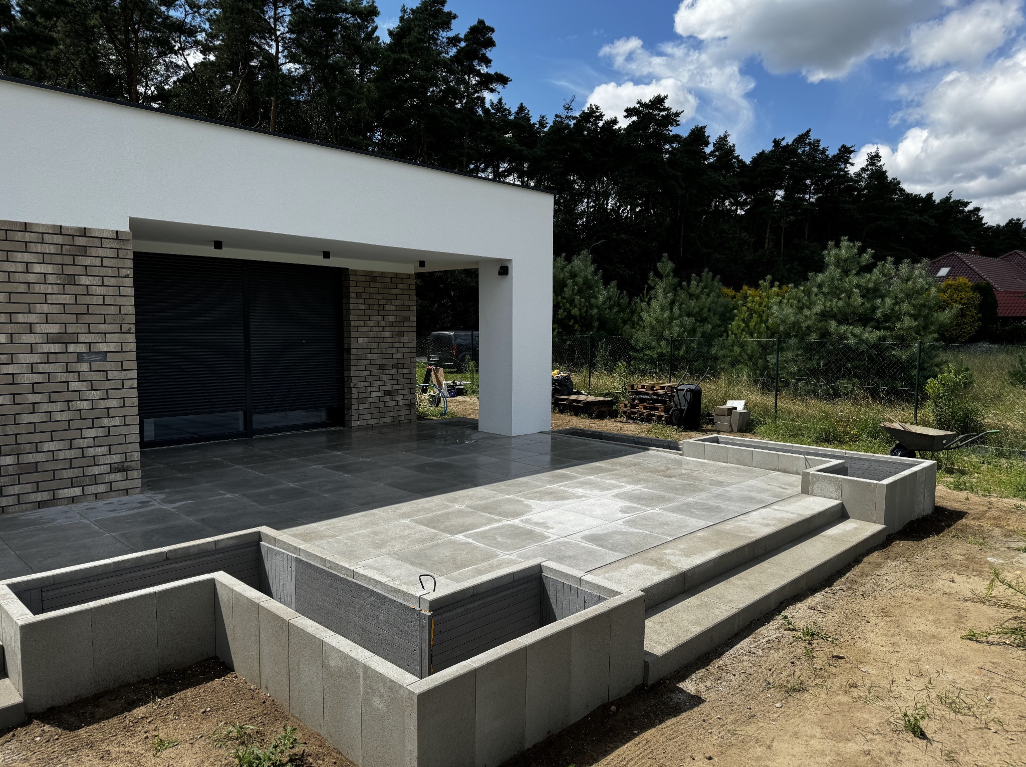 Nowoczesny taras z szarych płyt, częściowo wykończony, z widoczną konstrukcją pod rabaty. W tle dom z ceglaną elewacją i las.