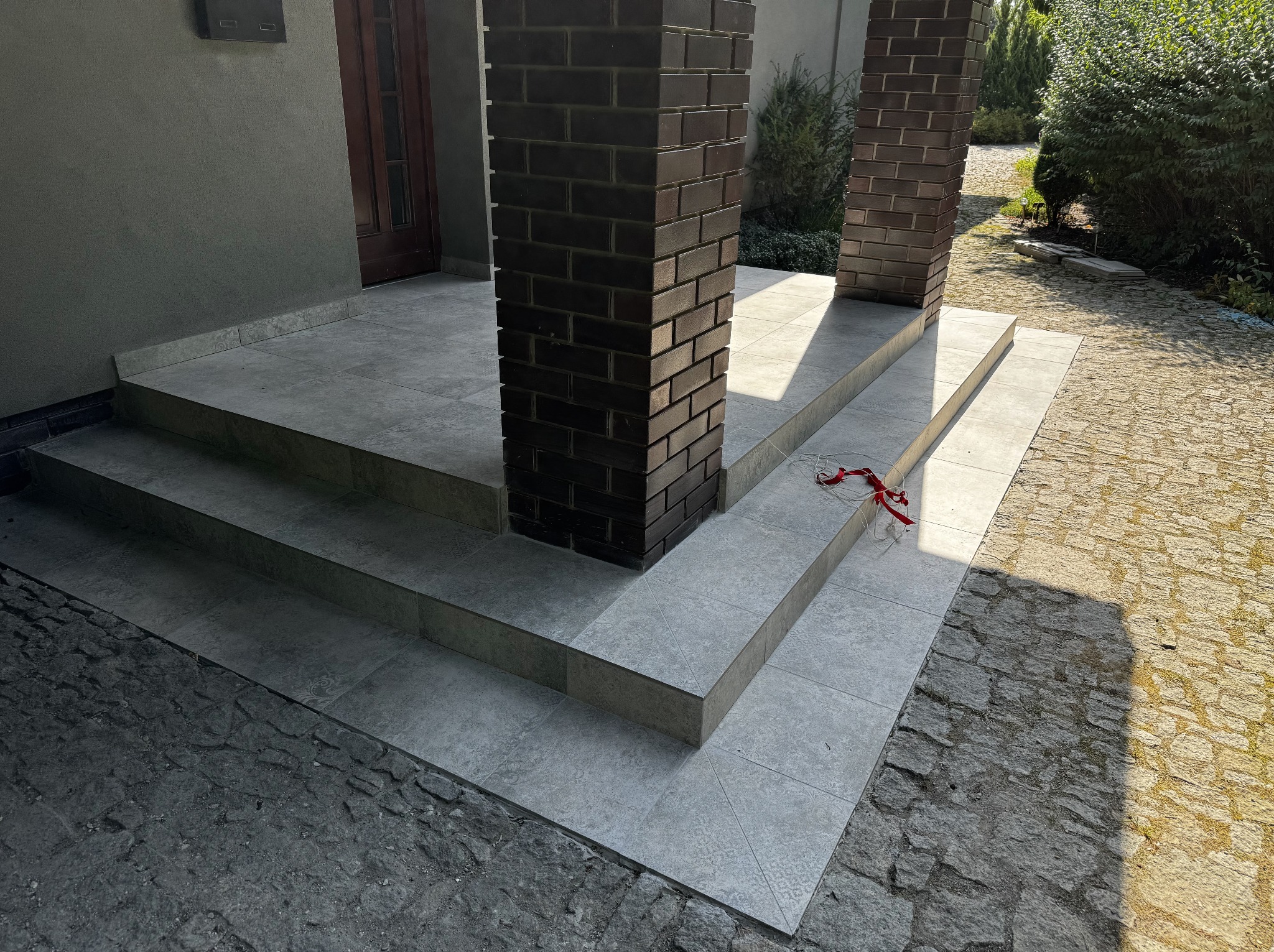 Wykończone płytkami schody wejściowe z ceglanymi filarami, prowadzące do drzwi. Na zewnątrz widoczna kostka brukowa i zieleń. Ujęcie z dołu, podkreślające detale.