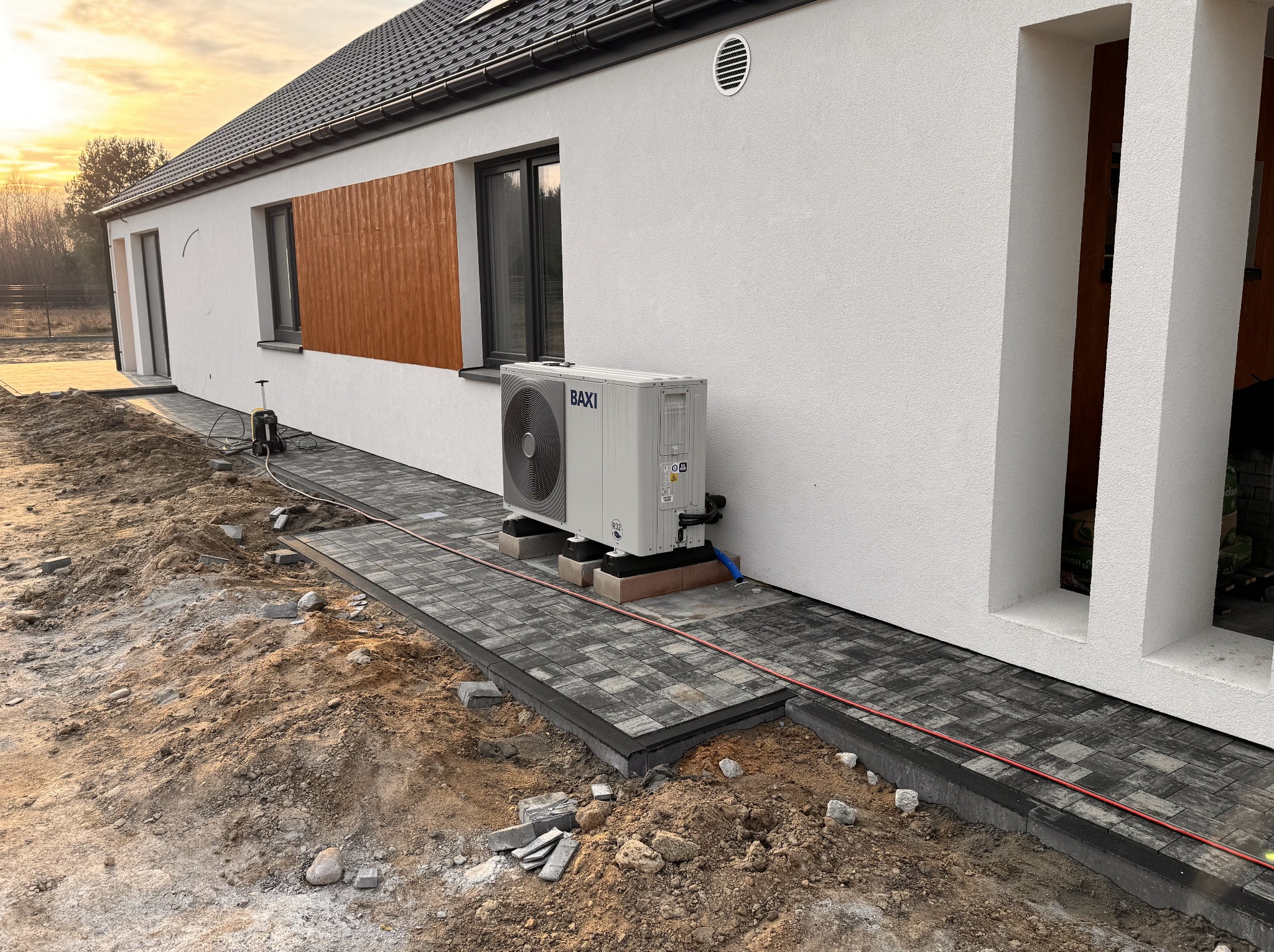 Fragment układanej ścieżki z kostki brukowej przy białym domu z klimatyzatorem. Widoczne narzędzia i elementy kostki do ułożenia. Częściowo zachmurzone niebo.