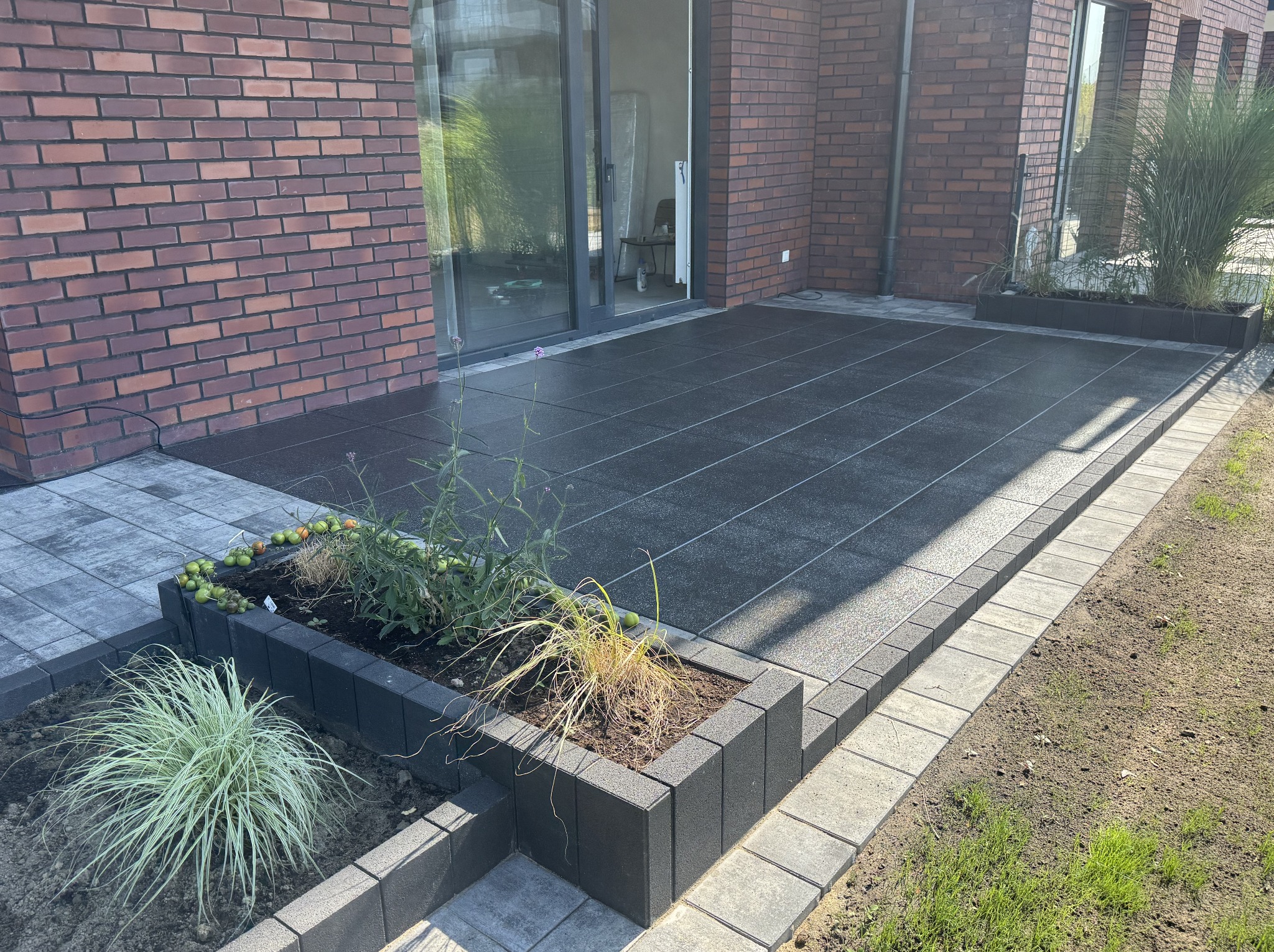 Nowoczesny taras z ciemnej kostki, obrzeża z jasnej kostki, donice z roślinami, ceglana ściana budynku w tle, przesuwne drzwi tarasowe.