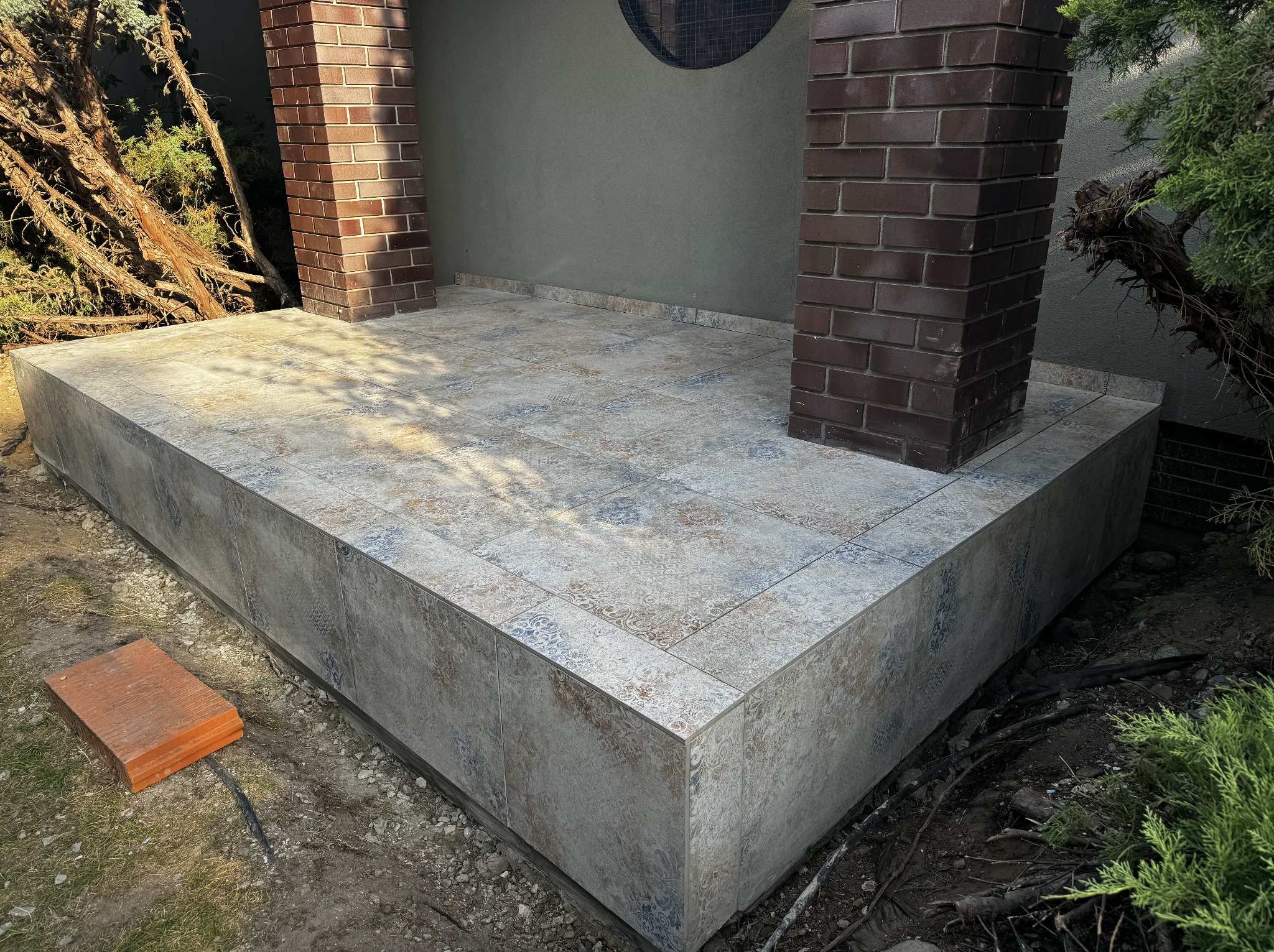 Wykończony taras z płytkami ceramicznymi w odcieniach beżu i błękitu, z ceglanymi filarami. Widoczne elementy ogrodu i pojedyncza płytka na ziemi.