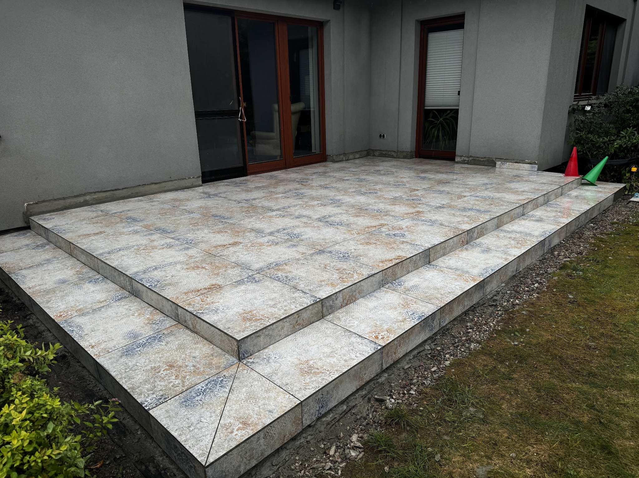 Wykończony taras z dwoma stopniami, wyłożony wzorzystymi płytkami ceramicznymi w odcieniach beżu i błękitu. Widoczne drzwi tarasowe i okno. Na trawie stoją dwa pachołki.