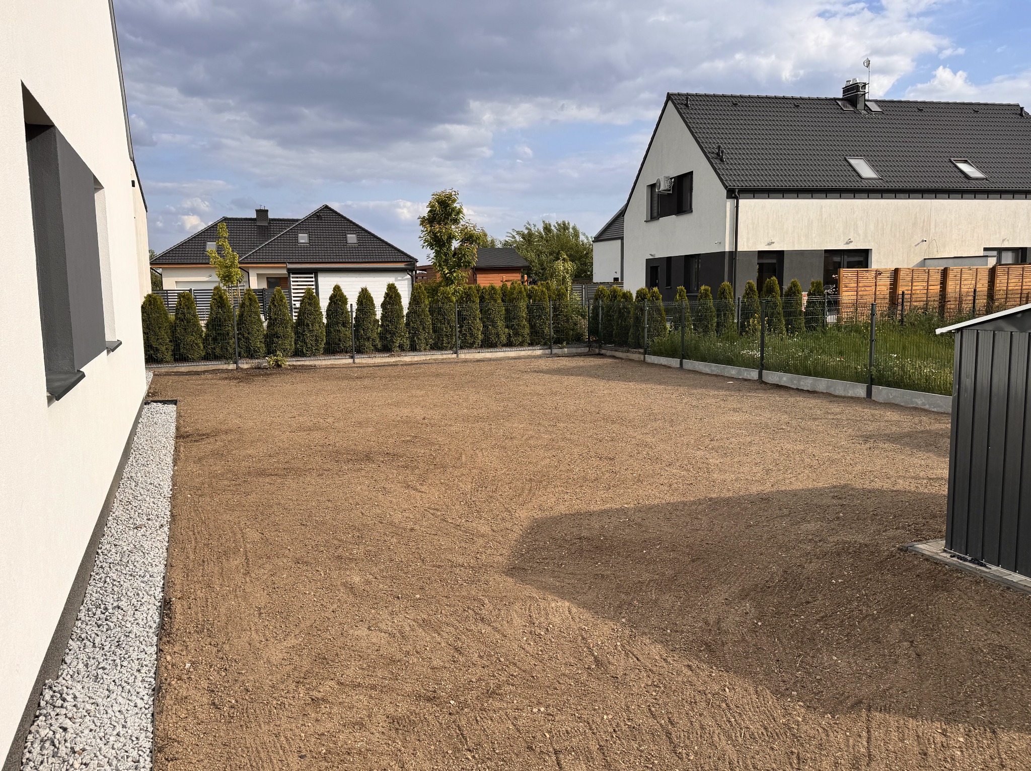 Przygotowany teren pod przyszły ogród w Poznaniu. W tle widoczne domy jednorodzinne i rząd zasadzonych drzewek oddzielających posesje.