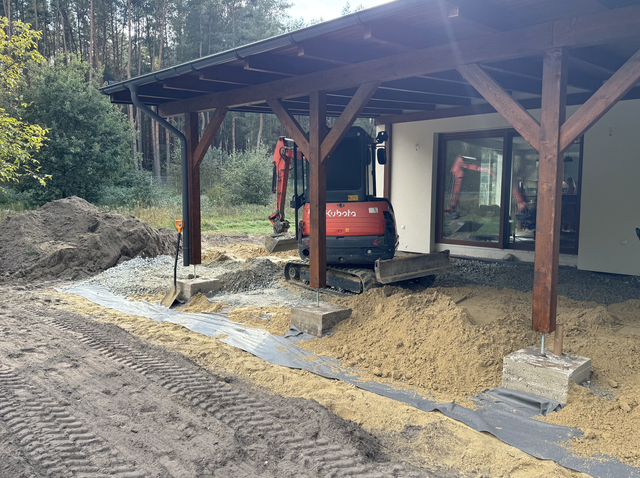Przygotowanie podłoża pod taras: mini koparka Kubota, piasek, geowłóknina i podsypka kamienna. Widoczne drewniane słupy z betonowymi podstawami.