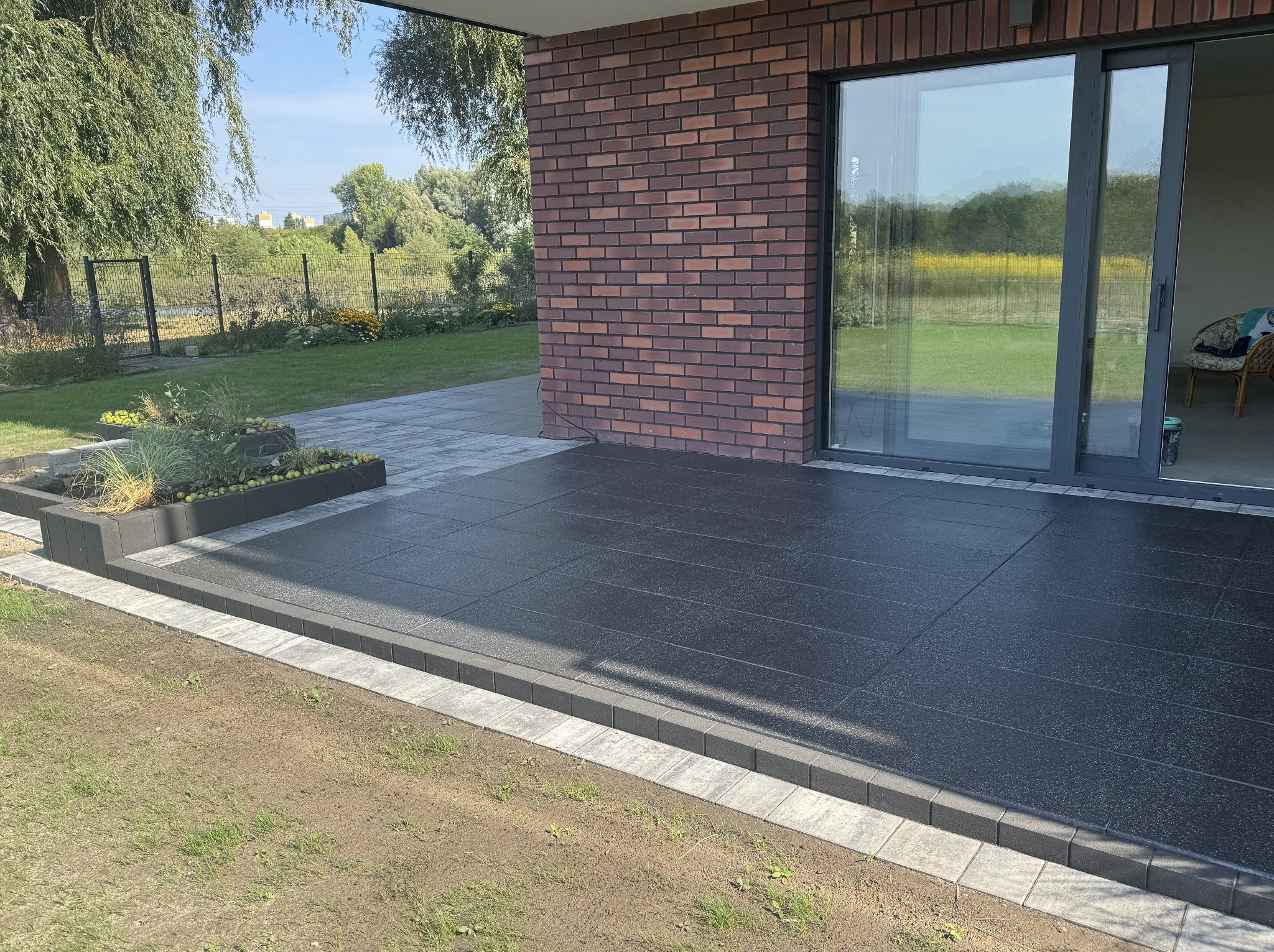Nowoczesny taras z ciemnych płyt, obrzeża z kostki, rabata z roślinami, ceglana ściana budynku i przesuwne drzwi tarasowe. Estetyczne wykończenie.