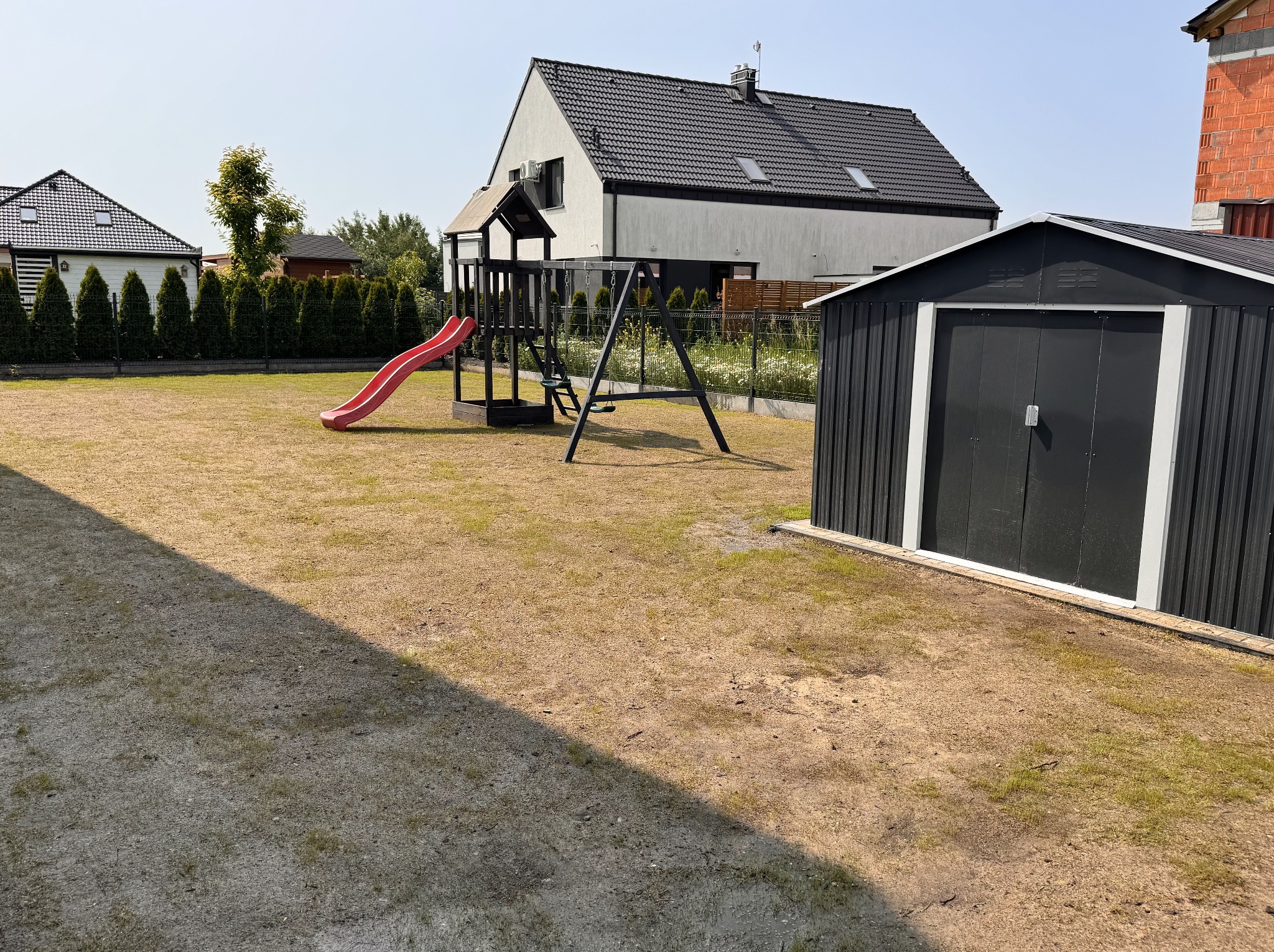 Przydomowy plac zabaw z huśtawką i zjeżdżalnią na trawniku, obok blaszany domek narzędziowy. W tle nowoczesny dom jednorodzinny z ciemną dachówką.