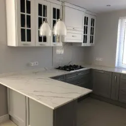 Nowoczesna kuchnia w odcieniach szarości i bieli z blatem o marmurowym wzorze, oświetlona wiszącymi lampami z przezroczystego szkła.