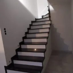 Nowoczesne schody o czarnych stopniach z białymi podstopnicami, widoczne niedokończone otwory na gniazdka elektryczne na ścianie.