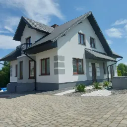 Dwupiętrowy dom z szarą dachówką i białymi ścianami, z szarymi akcentami, widoczny z poziomu brukowanego podjazdu, obok zaparkowany biały samochód dostawczy, w tle zielony ogród i błękitne niebo.