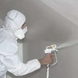 Pracownik w białym kombinezonie ochronnym aplikuje natryskowo białą substancję na skośną ścianę za pomocą pistoletu lakierniczego.