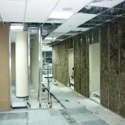 Montaż ścian działowych w biurze: metalowy szkielet, izolacja z wełny mineralnej, widoczne płyty kartonowo-gipsowe i drabina aluminiowa w tle.