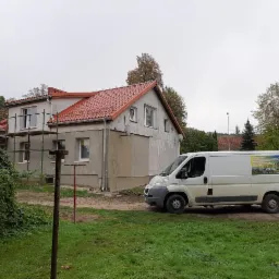 Dom z częściowo wymienionym dachem, widoczne rusztowania i samochód dostawczy z reklamą firmy, pochmurny dzień.