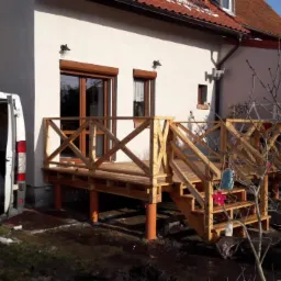 Drewniany taras z balustradą przy białym domu z czerwonym dachem, wsparty na pomarańczowych rurach, z widocznym samochodem dostawczym z reklamą firmy.