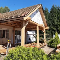 Wznoszenie drewnianej konstrukcji z fragmentami ceramicznej dachówki, widoczny laserowy niwelator na statywie i rusztowania przy budynku w tle.