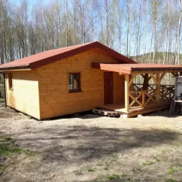 Nowoczesny, parterowy budynek z jasnego drewna z czerwonym dachem i zadaszonym tarasem z drewnianą konstrukcją kratową, na tle drzew bezlistnych.