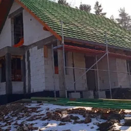 Niedokończony dom z bloczków z zielonym dachem, widoczne rusztowania i resztki śniegu na ziemi.