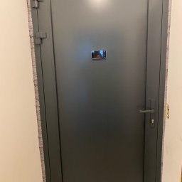 AluGlobal Spółka Cywilna Artur Pyła, Paulina Chemperek, Dawid Dubiel - Szare drzwi wejściowe z ościeżnicą, widoczne zawiasy i klamka, w centralnej części drzwi odbicie w telefonie komórkowym.