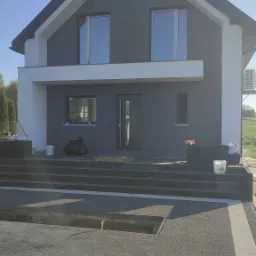 Nowoczesny dom jednorodzinny z szarą elewacją i białymi akcentami, widoczne okna z folią ochronną, teren przed domem w trakcie prac wykończeniowych.