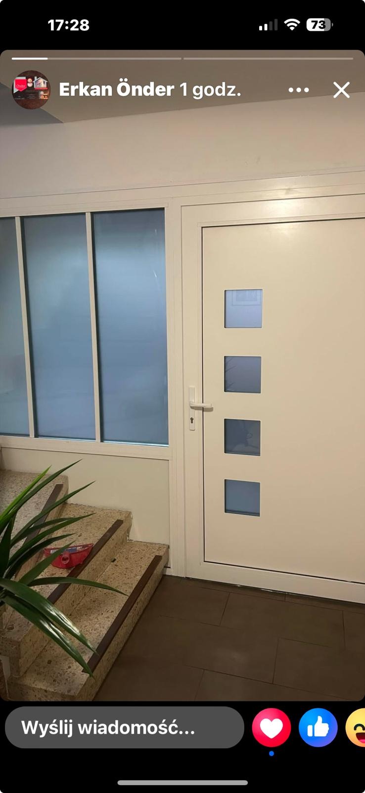 Białe drzwi wejściowe z czterema kwadratowymi przeszkleniami, po lewej stronie okno z matowym szkłem, schody z terazzo i roślina doniczkowa.