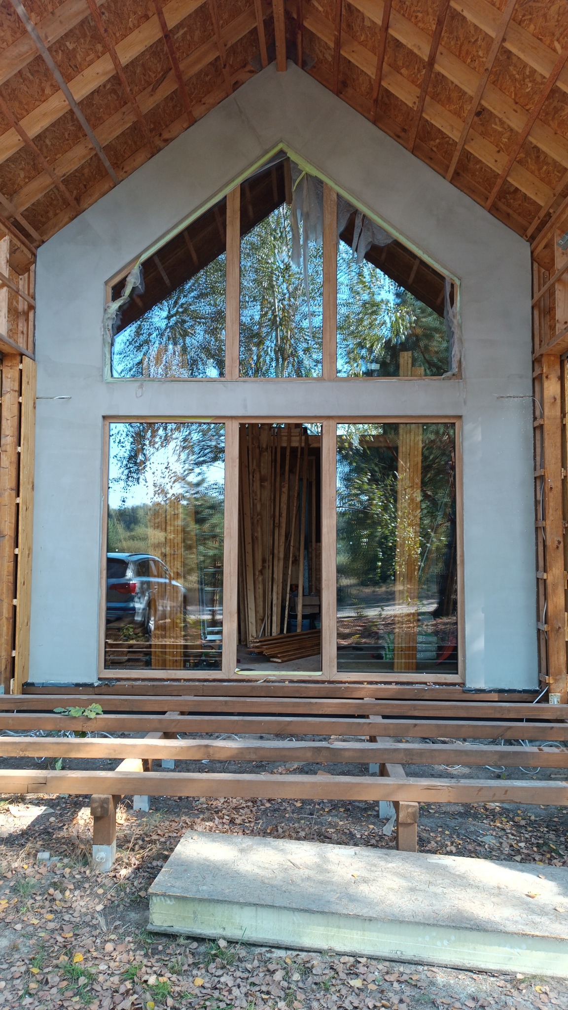 Montaż okien aluminiowych z drewnianą ramą w surowym stanie, widoczne rusztowanie pod podłogę i odbicie samochodu w szybie.