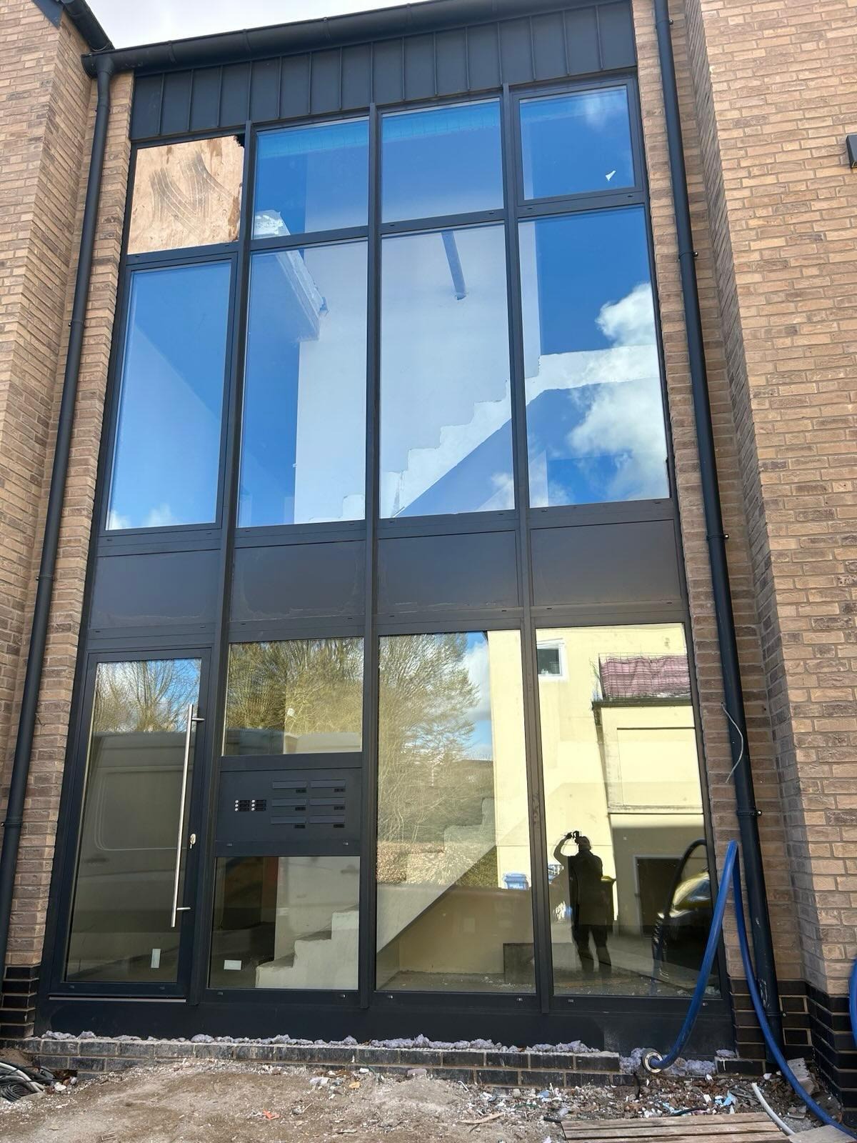 Duże przeszklenie w nowym budynku z cegły, z widoczną klatką schodową wewnątrz i odbiciem otoczenia w szybach, jedna z szyb zabita deskami.