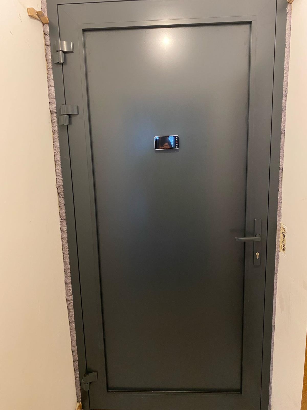 Szare drzwi wejściowe z ościeżnicą, widoczne zawiasy i klamka, w centralnej części drzwi odbicie w telefonie komórkowym.
