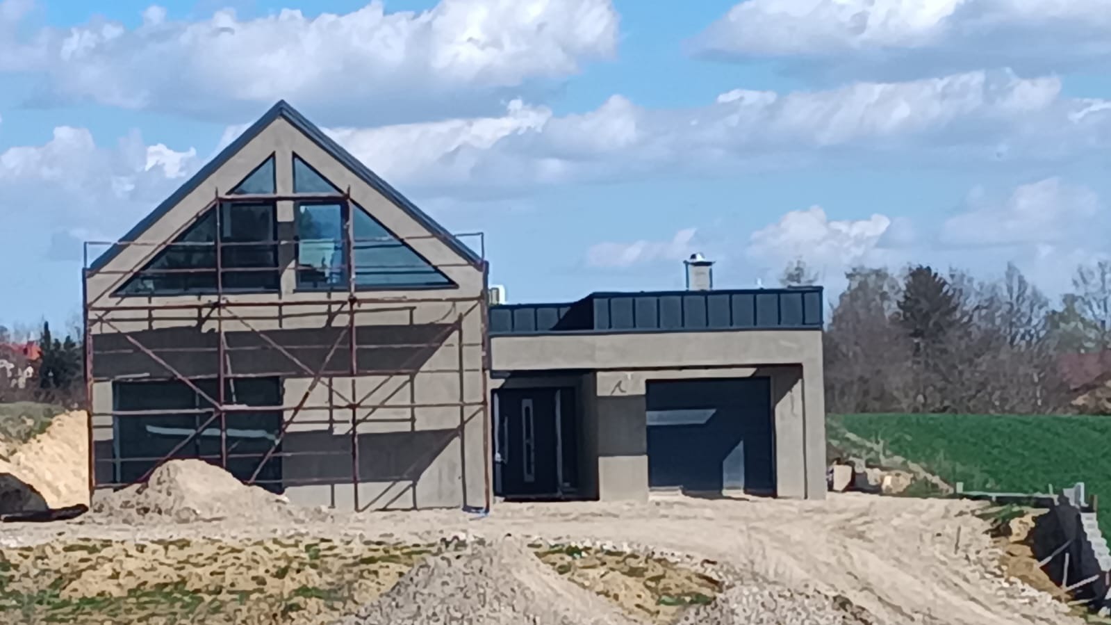 Dom w trakcie budowy z zamontowanymi oknami PCV o nietypowym trójkątnym kształcie, widoczne rusztowania i teren budowy.