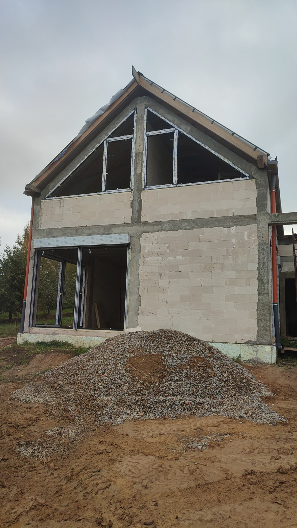 Budowa domu w stanie surowym otwartym, widoczne zamontowane ramy okienne, ściany z bloczków i pryzma żwiru przed budynkiem.