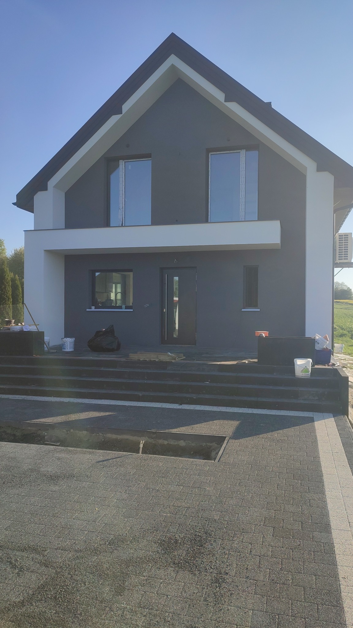 Nowoczesny dom jednorodzinny z szarą elewacją i białymi akcentami, widoczne okna z folią ochronną, teren przed domem w trakcie prac wykończeniowych.