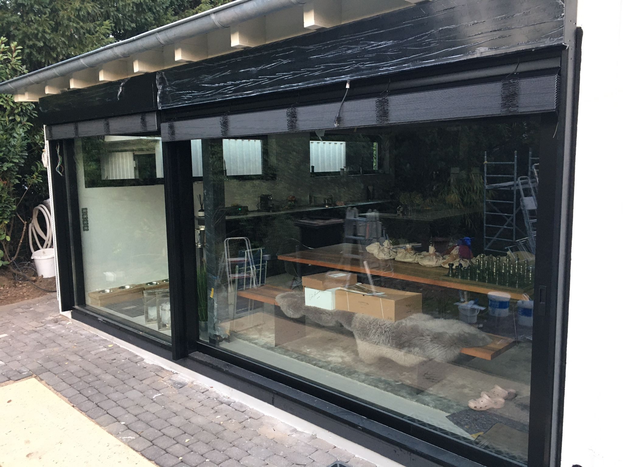 Duże przesuwne okno z czarną ramą i roletą zewnętrzną, widok na wnętrze z elementami dekoracyjnymi i narzędziami malarskimi