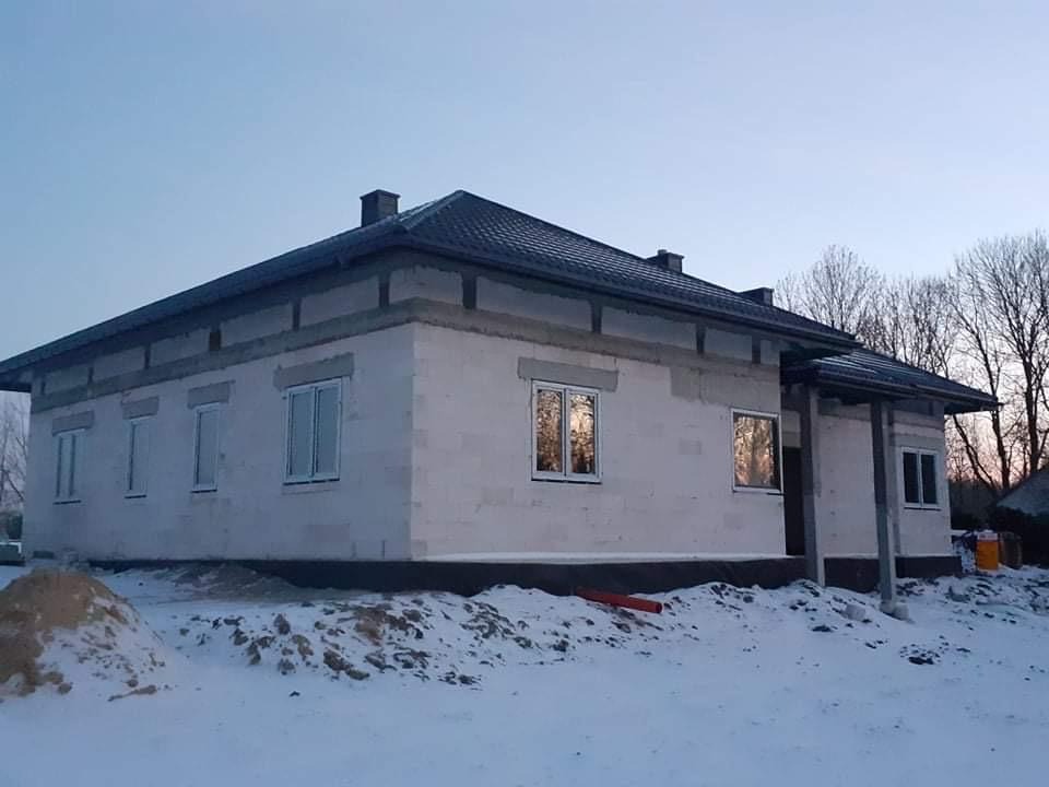 Surowy stan domu jednorodzinnego w zimowej scenerii, widoczne białe ramy okienne, ściany z pustaków i zasypane śniegiem podwórko.