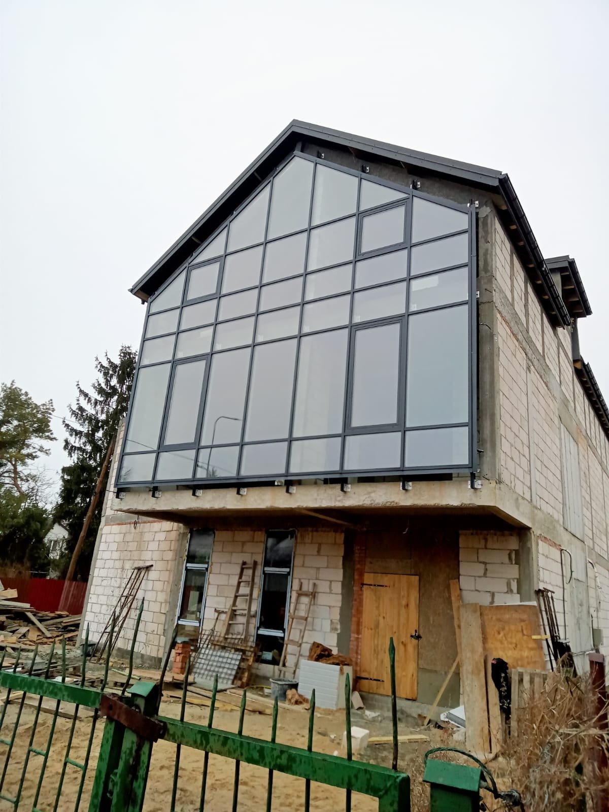 Dom w trakcie budowy z zamontowaną dużą ścianą okienną z czarnymi ramami, widoczne rusztowania i materiały budowlane.