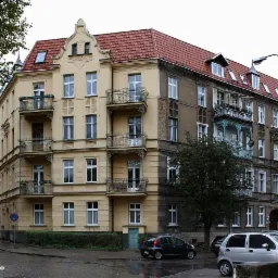 Kamienica narożna z żółtą elewacją po lewej i szarą elewacją po prawej, z balkonami i czerwonym dachem, widok z poziomu ulicy na mokrym chodniku, zaparkowane samochody.