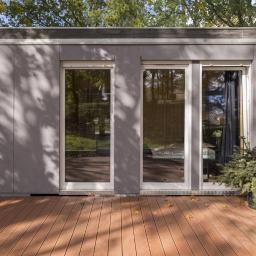 MagaHomes - Nowoczesny, modułowy budynek z szarej elewacji z trzema wysokimi oknami, usytuowany na drewnianym tarasie, otoczony zielenią drzew.