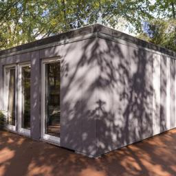 MagaHomes - Nowoczesny, parterowy budynek o szarej elewacji z dużymi oknami, otoczony zielenią drzew i drewnianym tarasem. Na tarasie widoczne małe drzewka iglaste w czarnych donicach.