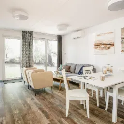 Jasne wnętrze domu z drewnopodobną podłogą, beżowymi fotelami i białym stołem jadalnym, widok na ogród przez duże okna.